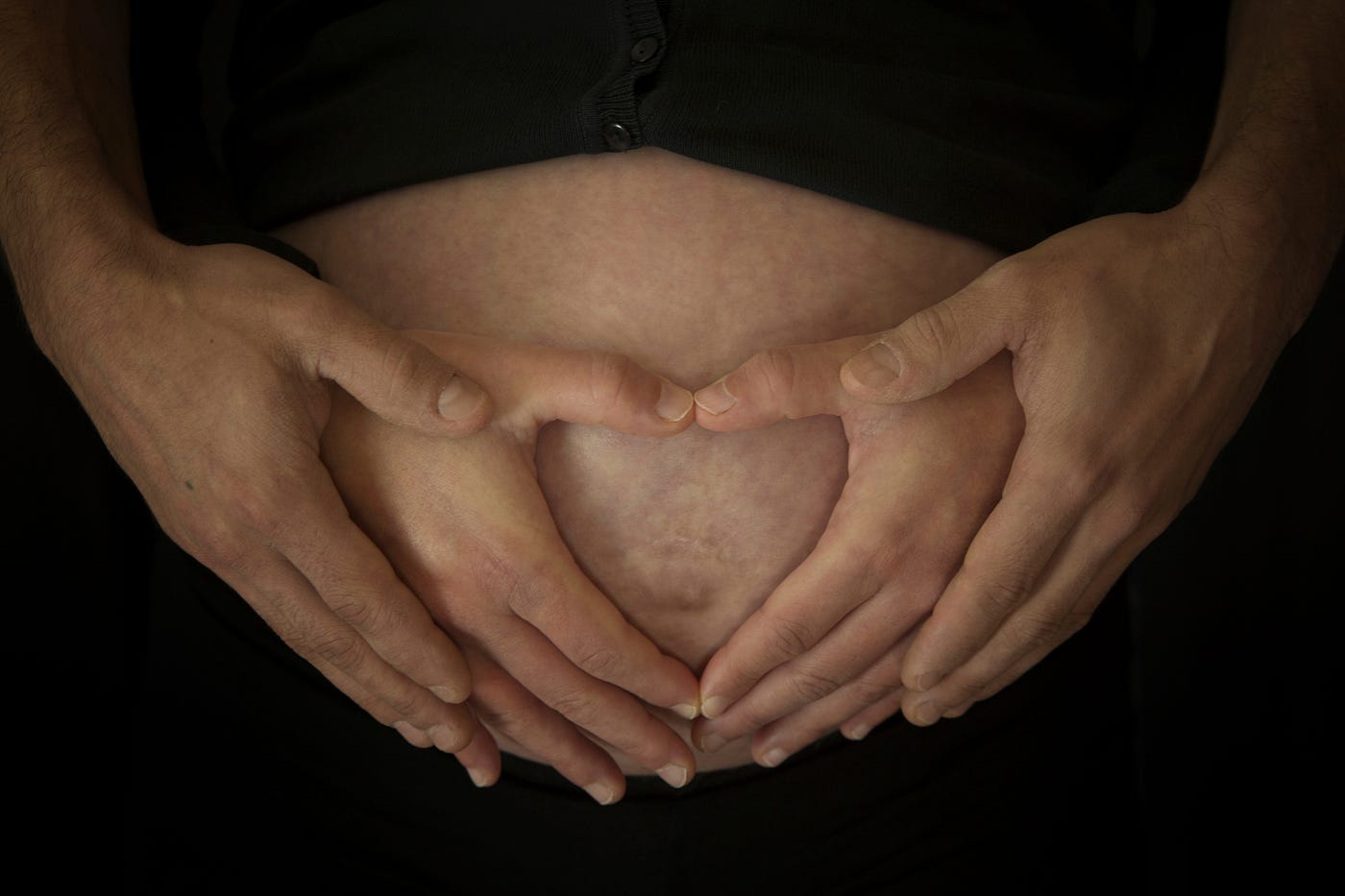 A close-up of a pregnant belly with two pairs of hands forming a heart shape over it, symbolizing love and anticipation for the baby.