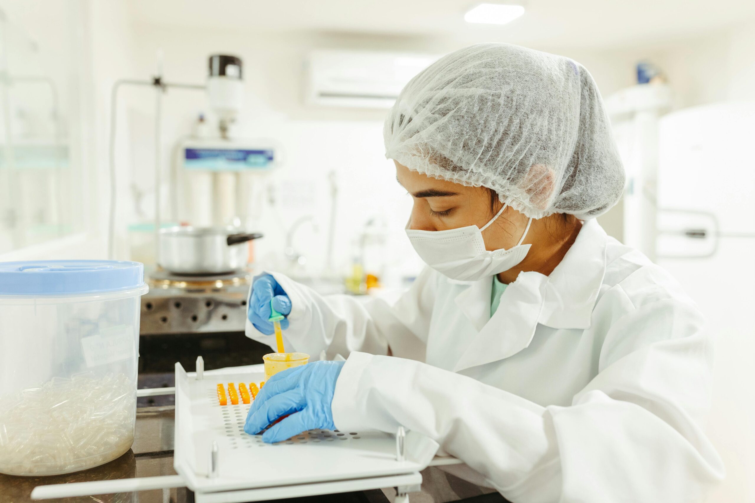 A laboratory technician wearing a hairnet, mask, and gloves works with samples in a sterile laboratory environment, focusing on precise measurements and careful handling.