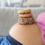 Pregnant woman with donuts on belly. Cravings of desserts and sweets during pregnancy, Girl eating unhealthy pastries on baby bump for gestational diabetes.