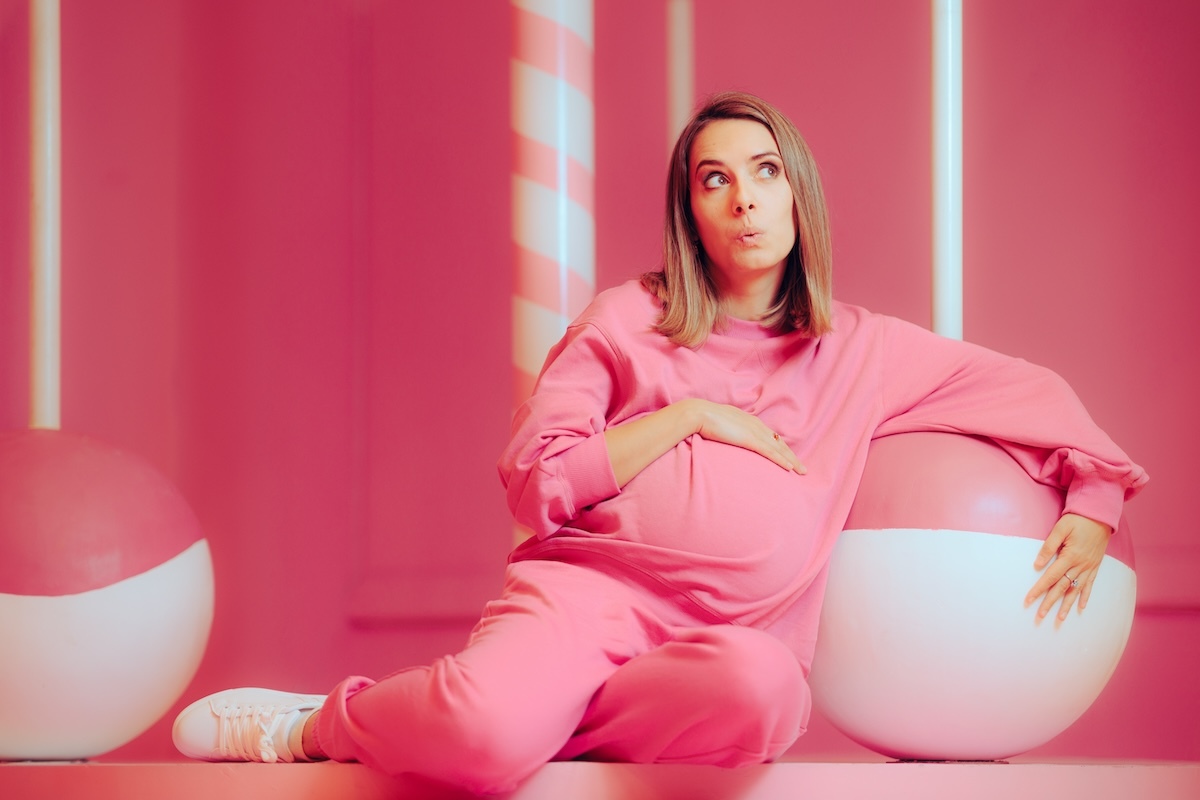 A pregnant woman in a pink outfit sitting in a pink room, resting against a large pink and white ball.