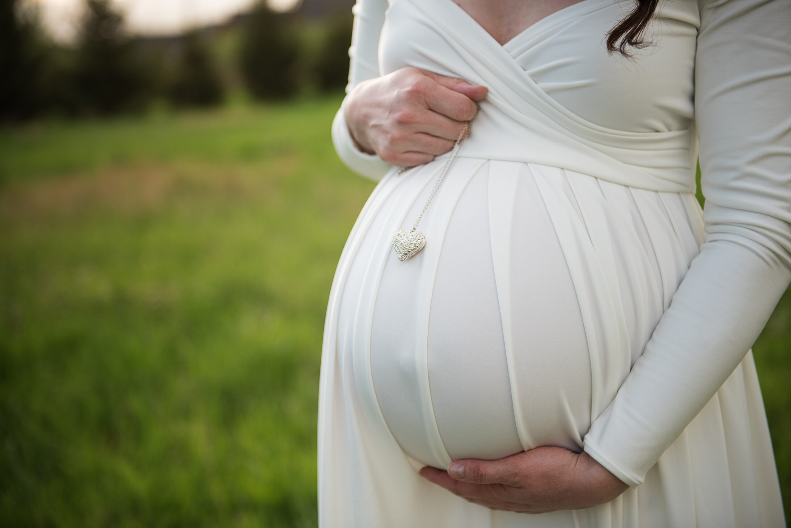 A close-up of a pregnant woman in a white, long-sleeved dress, holding her belly with both hands. A heart-shaped pendant rests on her round abdomen, and she stands outdoors in a grassy field.