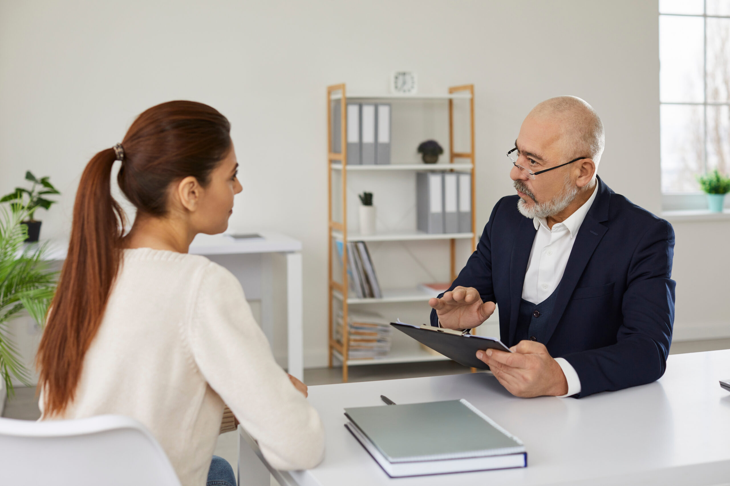 A woman with a ponytail sits across a desk from an older man in a suit, who is holding a clipboard and appears to be explaining something to her. They are in a modern, well-lit office with shelves and plants in the background.