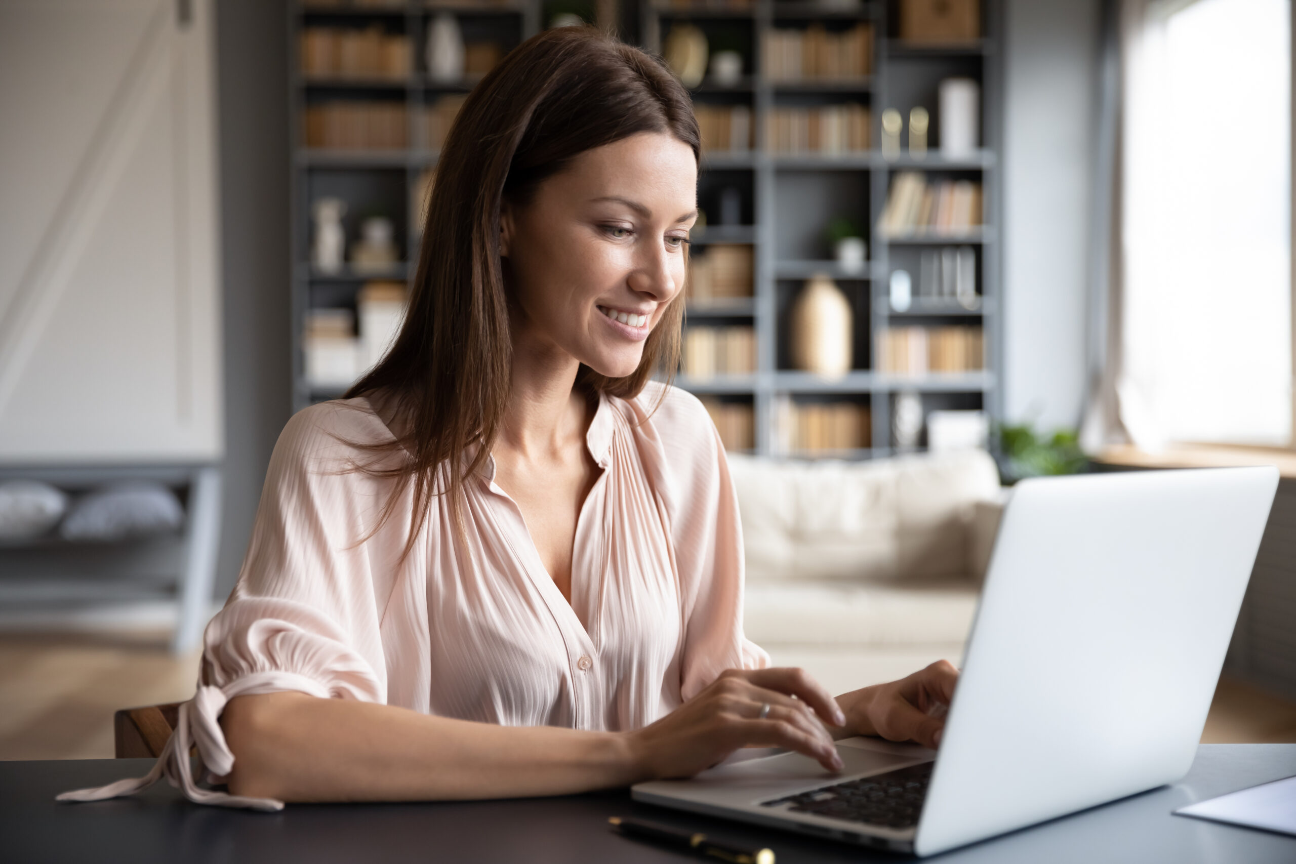 A woman smiling while looking at a laptop in a cozy home office with bookshelves in the background.