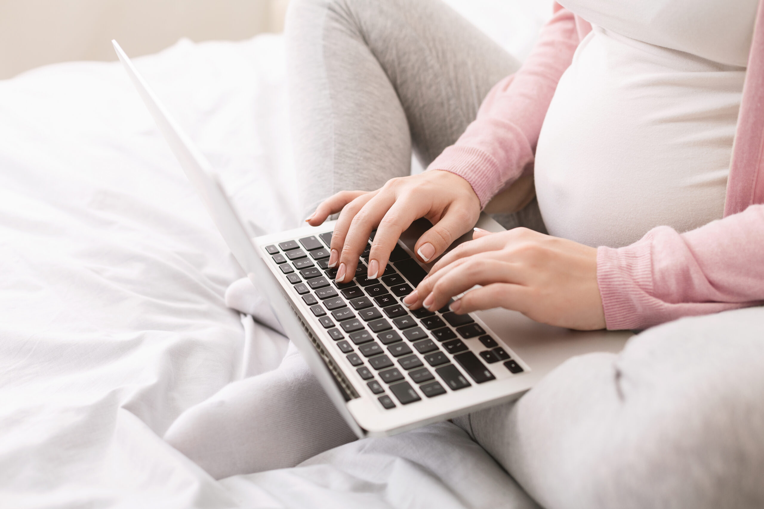 A pregnant woman sitting cross-legged on a bed, typing on a laptop, with the focus on her hands and the laptop.