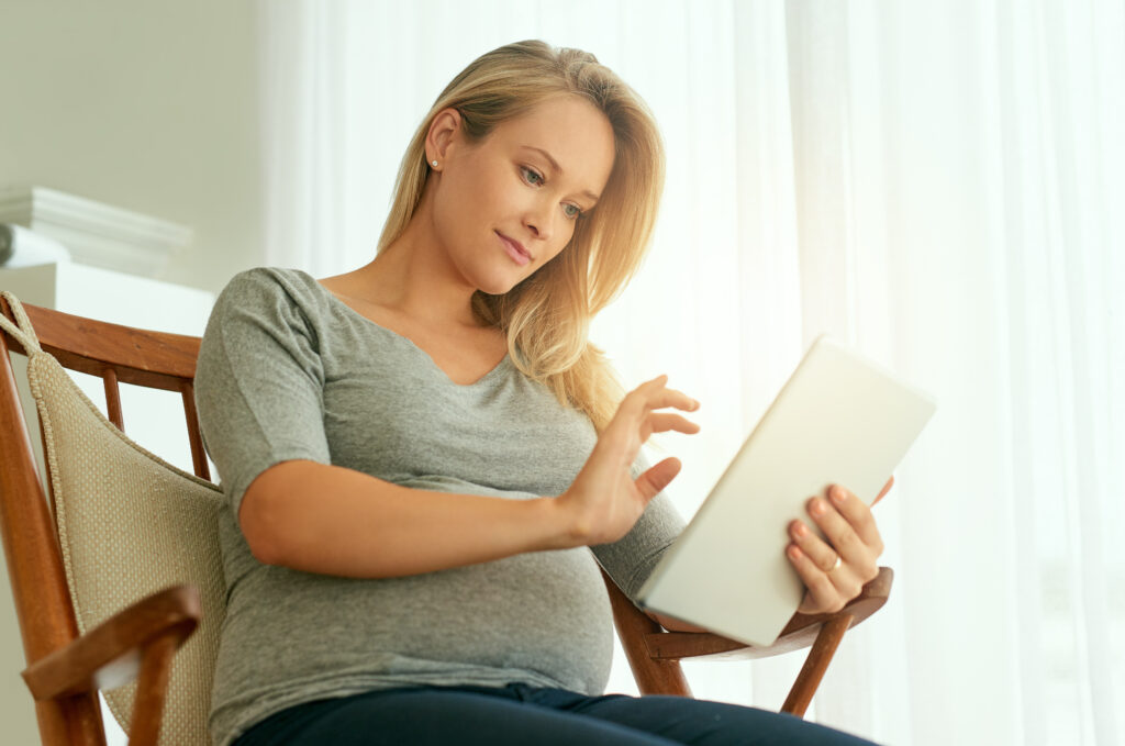 A pregnant woman sitting comfortably in a chair, using a tablet while gently resting her hand on her belly.