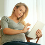 A pregnant woman sitting comfortably in a chair, using a tablet while gently resting her hand on her belly.