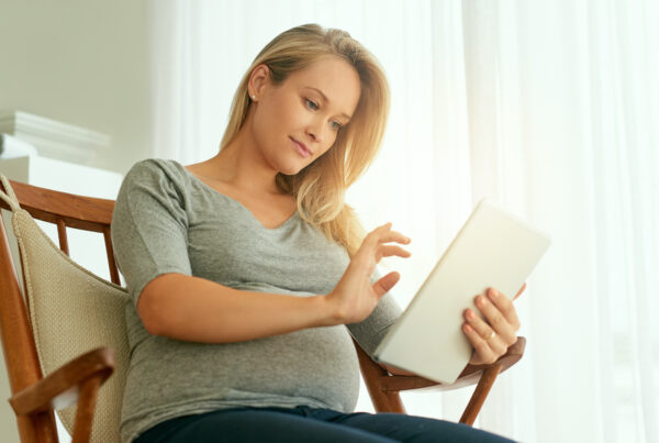 A pregnant woman sitting comfortably in a chair, using a tablet while gently resting her hand on her belly.