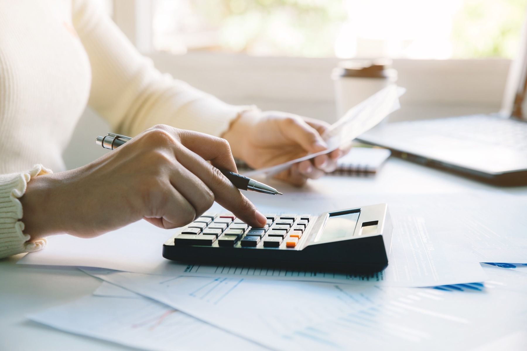 A person in a light-colored sweater uses a calculator while holding a paper, surrounded by financial documents and a laptop on a brightly lit desk.