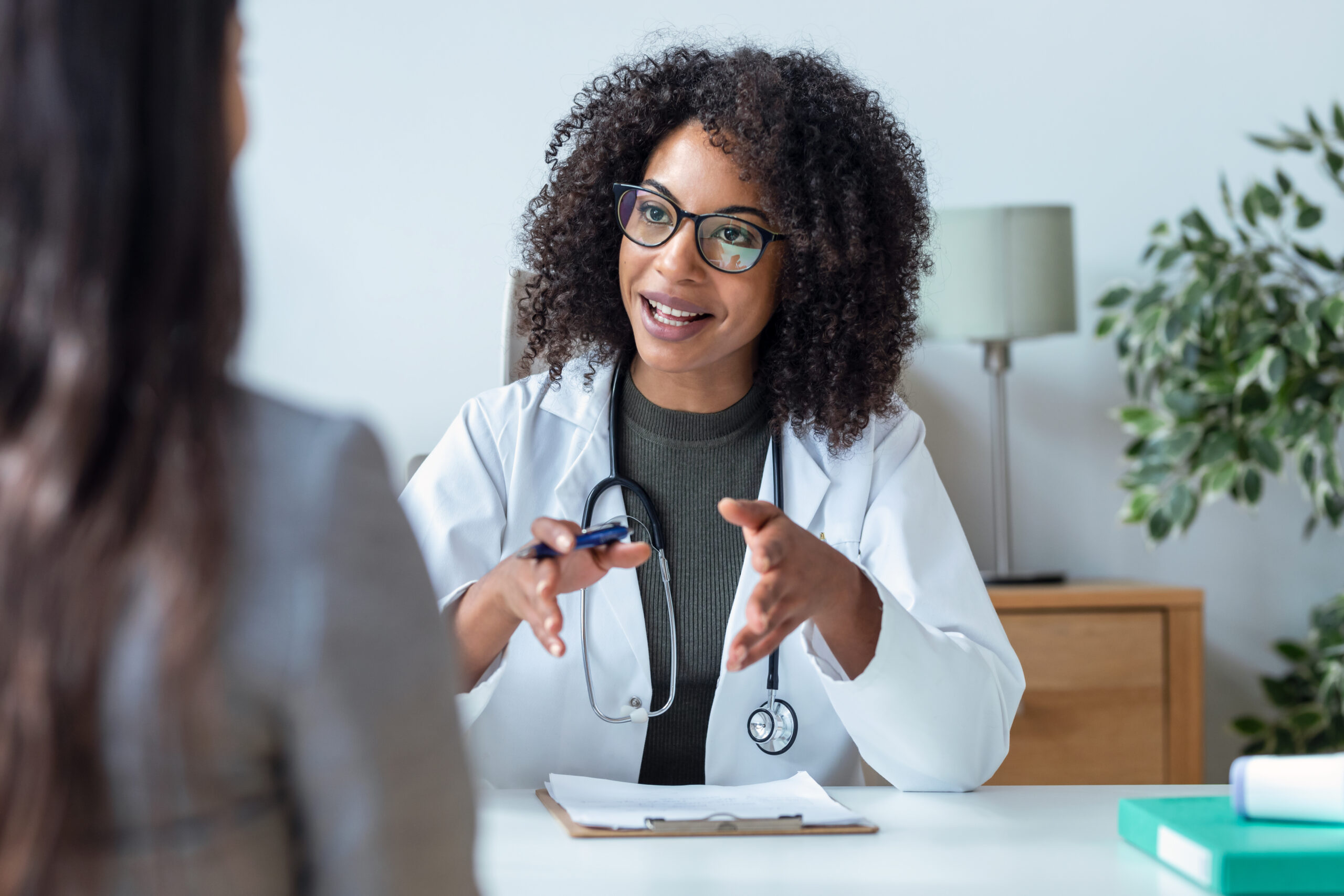 A female doctor with curly hair and glasses is seated at a desk, smiling and gesturing as she speaks to a patient. She has a stethoscope around her neck, and a clipboard with papers is in front of her.