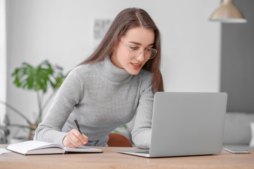 A young woman wearing glasses and a gray turtleneck sweater is studying at her desk, taking notes in a notebook while looking at a laptop in a bright and modern room.