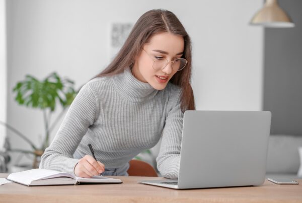 A young woman wearing glasses and a gray turtleneck sweater is studying at her desk, taking notes in a notebook while looking at a laptop in a bright and modern room.