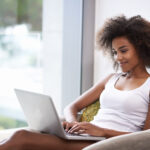 A woman with curly hair is seated in a comfortable chair near a window, smiling while using a laptop on her lap. She is wearing a white tank top and appears relaxed in a bright, serene indoor space.