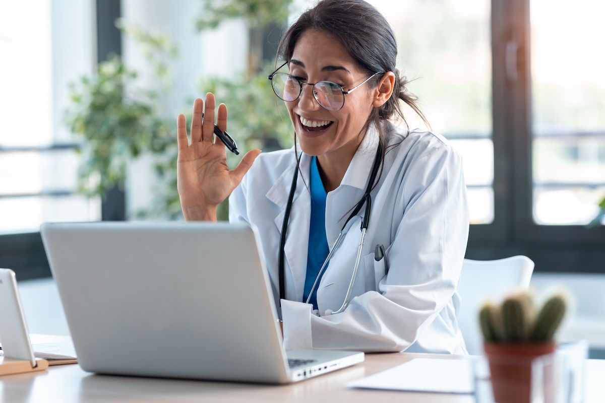 A female doctor wearing a white coat and glasses is smiling and waving while on a video call using a laptop. She has a stethoscope around her neck, and the background includes green plants and bright windows.