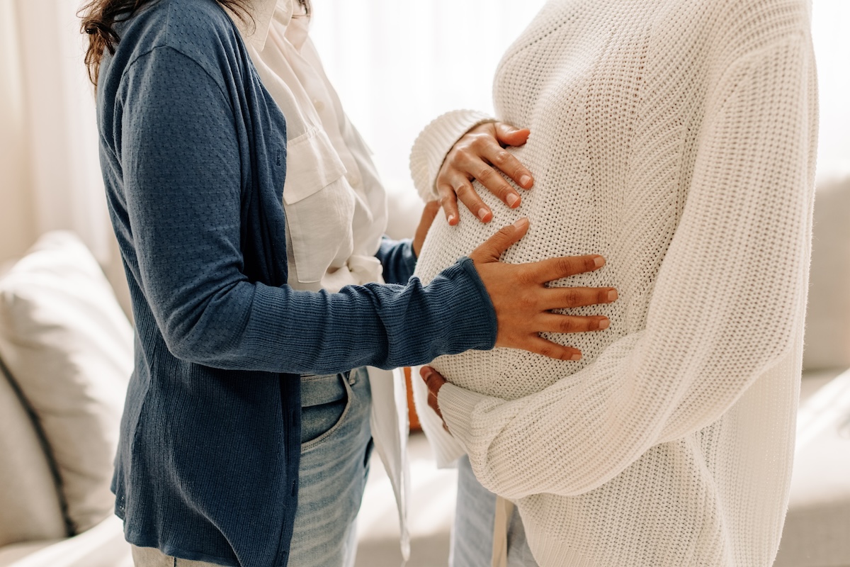 Two people interacting; one is touching the pregnant belly of the other. The person with the belly is wearing a white knit sweater, while the other wears a blue cardigan. The setting is a cozy, softly lit room.