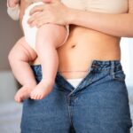 A woman holding a baby in a diaper against her hip. Her jeans are unbuttoned, revealing a faint scar on her lower abdomen, possibly from a recent cesarean section.