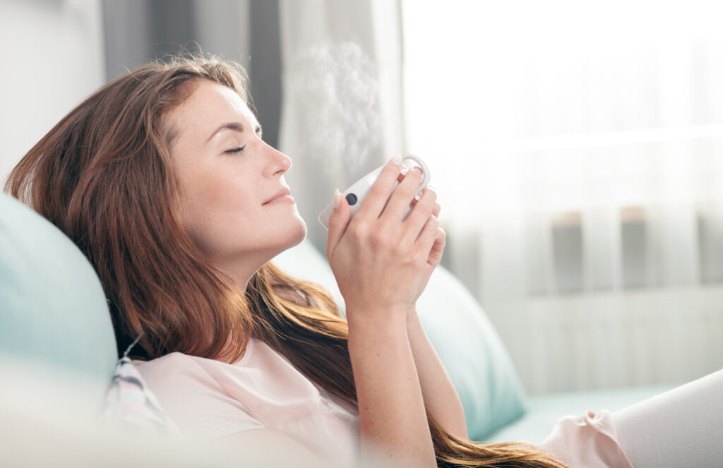 A relaxed woman with long hair sits on a couch, eyes closed, savoring the warmth and aroma of a steaming cup of coffee in a bright and cozy room.