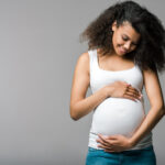 A smiling pregnant woman in a white tank top and jeans gently cradles her belly with both hands, standing against a plain gray background.