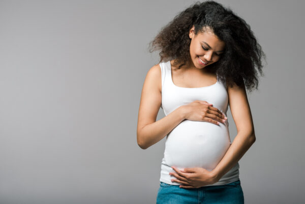 A smiling pregnant woman in a white tank top and jeans gently cradles her belly with both hands, standing against a plain gray background.