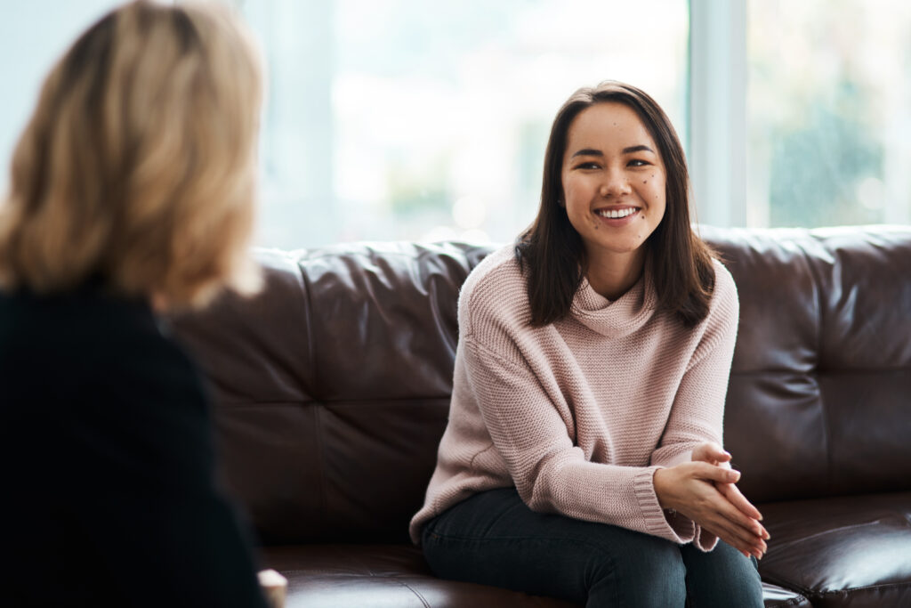 A cheerful young woman in a pink sweater sits on a brown leather couch, smiling warmly during a conversation in a bright, naturally lit room.