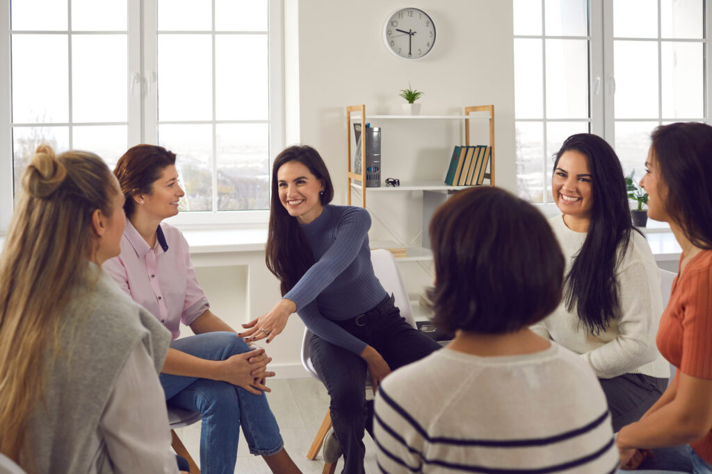 A group of women sits in a circle, engaged in a lively and supportive discussion in a bright room with large windows and minimal decor.