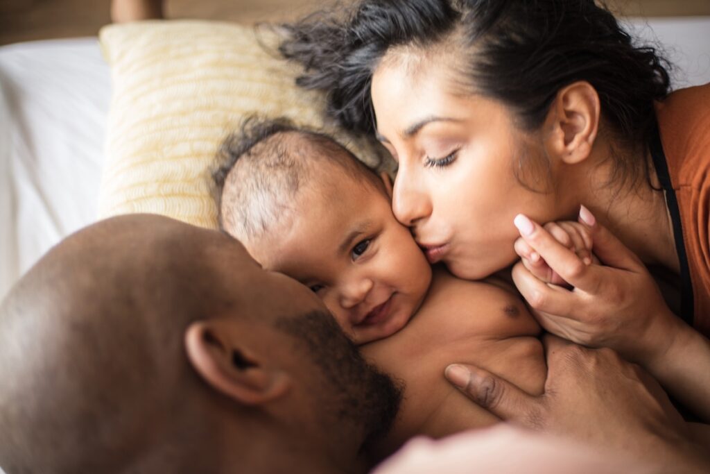 A baby lying between two adults on a bed, with one adult kissing the baby's cheek and the other holding the baby gently. The baby is smiling slightly, surrounded by warmth and affection.