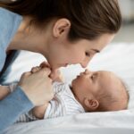 A woman in a blue shirt leaning close to a baby lying on a bed, holding the baby’s hands. They are making eye contact, with the woman smiling softly.