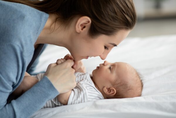 A woman in a blue shirt leaning close to a baby lying on a bed, holding the baby’s hands. They are making eye contact, with the woman smiling softly.