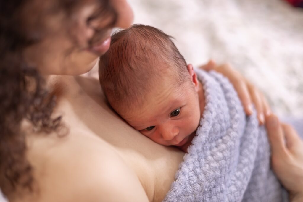 A newborn baby resting on an adult's chest, wrapped in a soft blue blanket. The baby looks calm and is snuggled closely for warmth and comfort.