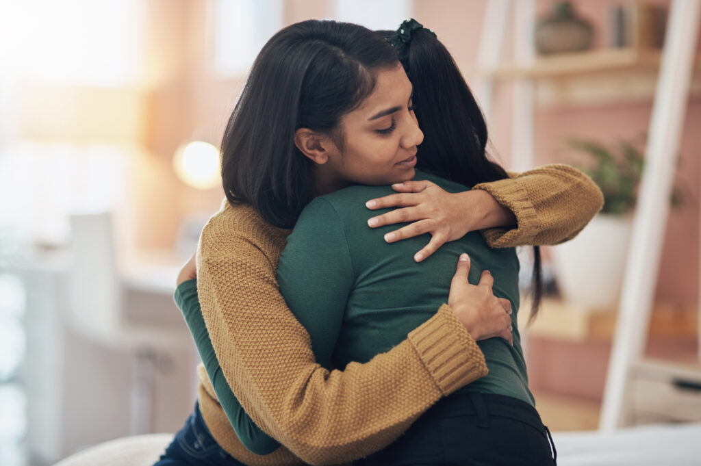  Two women embracing in a comforting hug in a warmly lit and cozy room, conveying support and empathy.