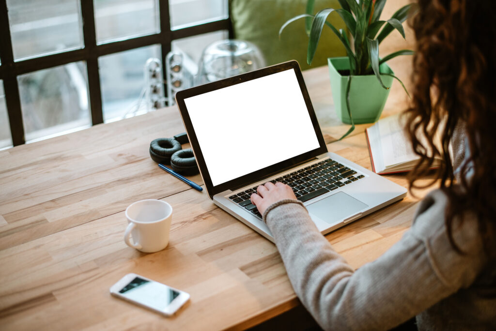 A person working on a laptop with a blank white screen, sitting at a wooden desk with a cup, headphones, a plant and a phone nearby. Natural light streams through large windows in the background.