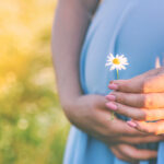 Close-up of a pregnant woman in a light blue dress holding a small daisy flower in her hand, standing in a sunlit field with a soft, blurred background.