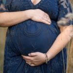 Pregnant woman in a navy lace dress cradling her baby bump with both hands, wearing colorful beaded bracelets and a ring, standing in an outdoor setting surrounded by tall grass.