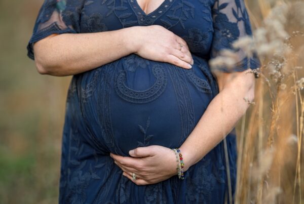 Pregnant woman in a navy lace dress cradling her baby bump with both hands, wearing colorful beaded bracelets and a ring, standing in an outdoor setting surrounded by tall grass.