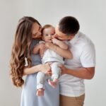 Loving parents holding their baby together, with the mother smiling down at the child and the father kissing the baby's cheek.