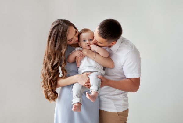 Loving parents holding their baby together, with the mother smiling down at the child and the father kissing the baby's cheek.