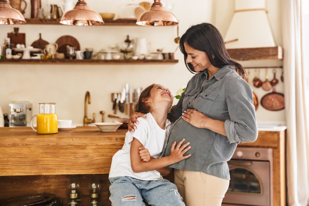 A surrogate mother smiles warmly as her daughter hugs her and looks up at her lovingly in a homey kitchen setting.