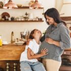 A surrogate mother smiles warmly as her daughter hugs her and looks up at her lovingly in a homey kitchen setting.