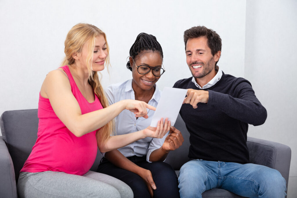 A surrogate mother in a pink tank top sits on a couch with the baby's intended parents, looking at a document.