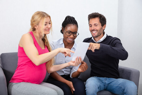 A surrogate mother in a pink tank top sits on a couch with the baby's intended parents, looking at a document.