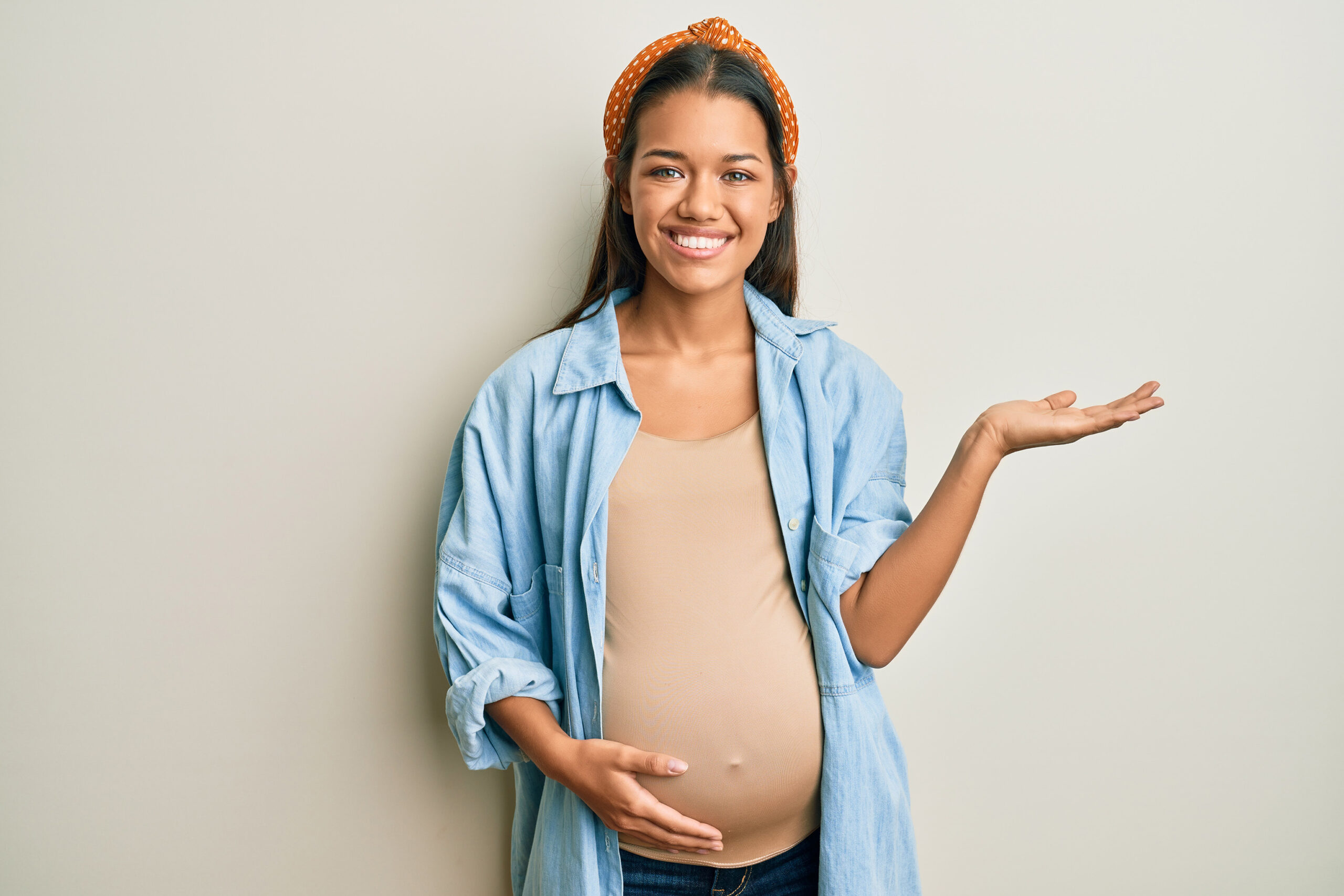 A smiling pregnant woman wearing a light denim shirt and an orange headband, standing against a plain background with one hand on her belly and the other gesturing to the side.