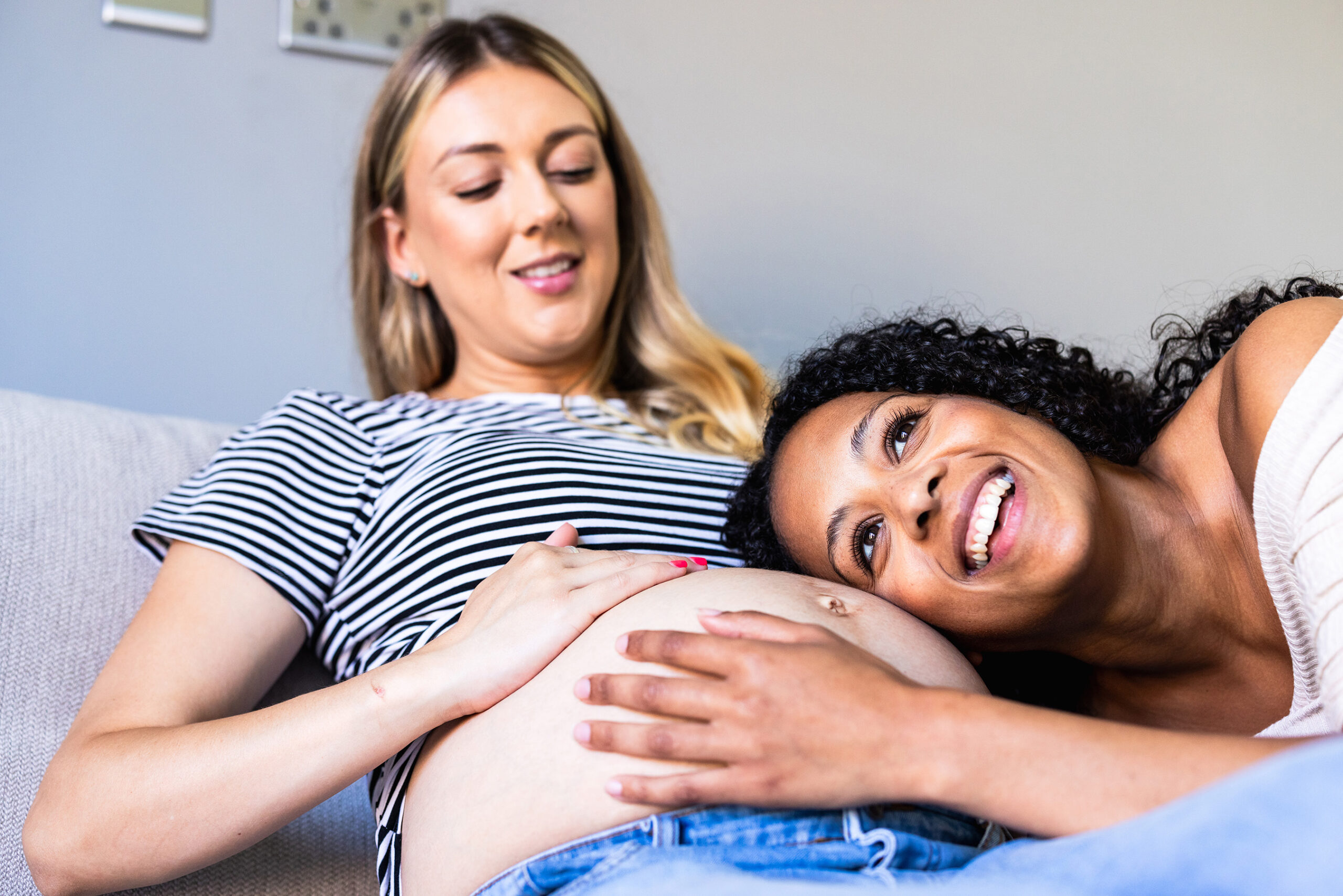 A surrogate mother relaxing on a couch while her friend smiles and rests her head on the surrogate's pregnant belly, sharing a joyful and supportive moment together.