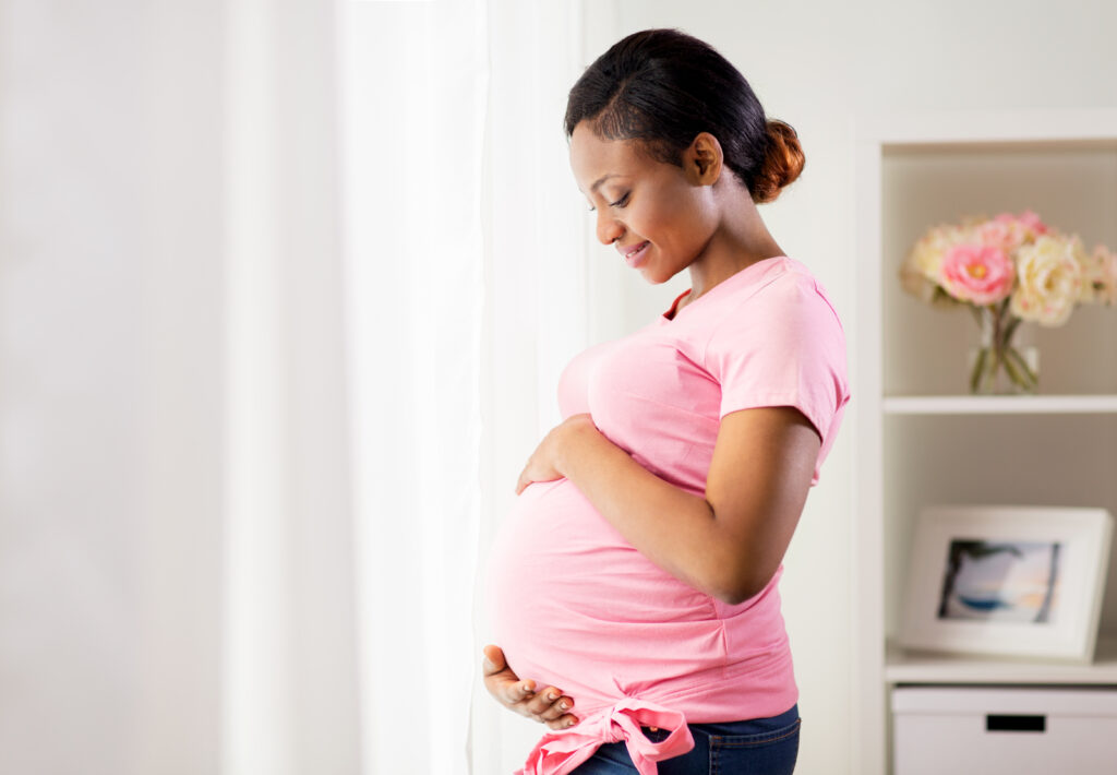 A surrogate mother in a pink shirt smiling and holding her belly while standing near a bright window.