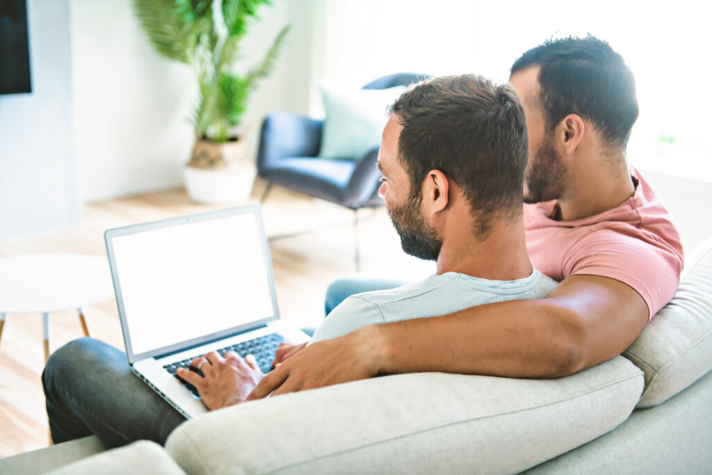 A same-sex male couple sitting on a couch, looking at a laptop together in a bright living room.