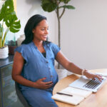 Pregnant woman smiling while using a laptop at a desk, researching surrogacy laws in a bright, modern home office.