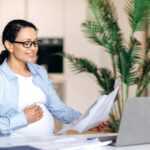 Pregnant woman working at a desk, smiling while reading papers in front of a laptop.