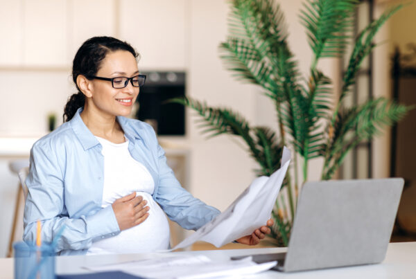 Pregnant woman working at a desk, smiling while reading papers in front of a laptop.