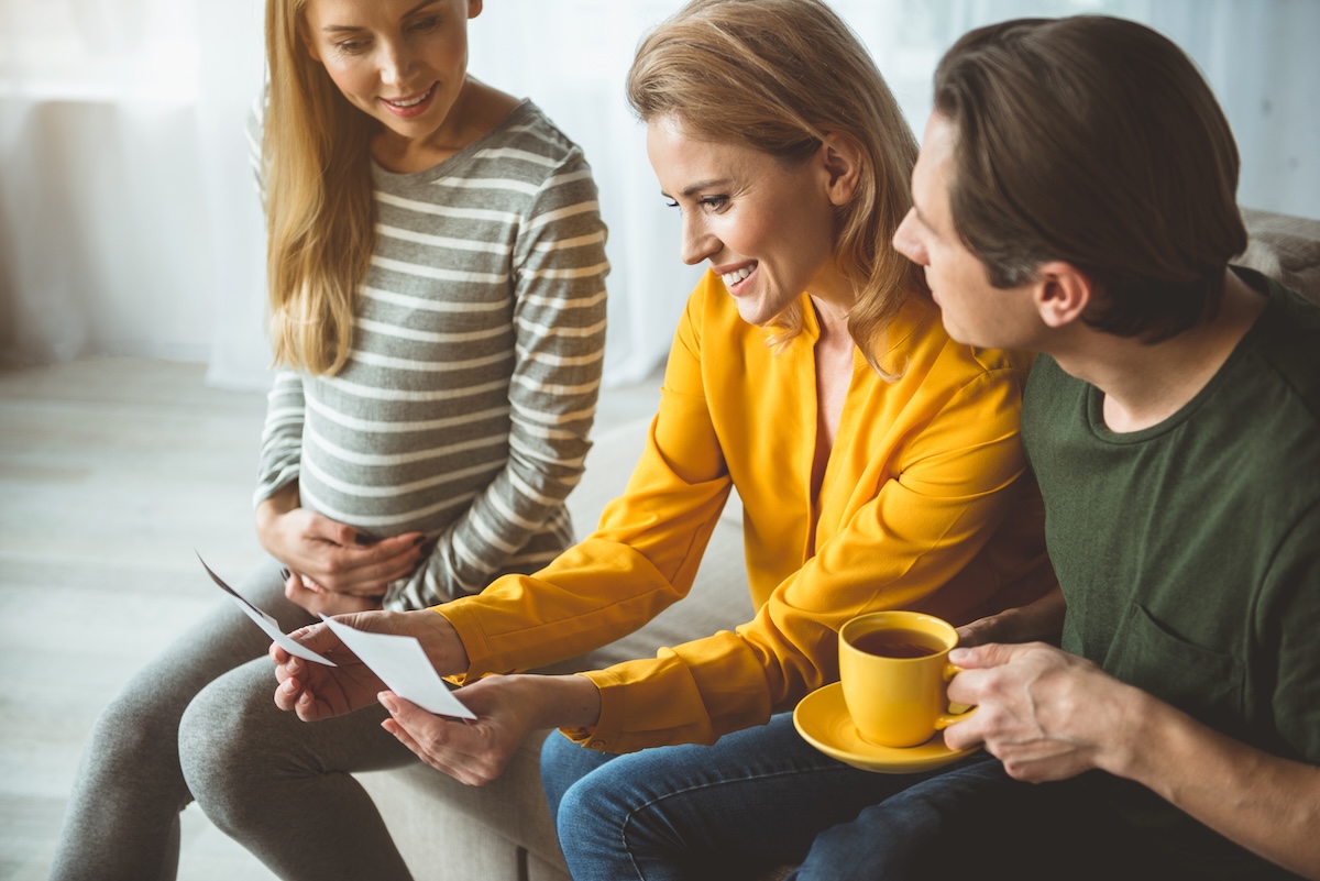 A surrogate mother sits next to her intended parents as they smile and look at ultrasound images together in a bright living room.