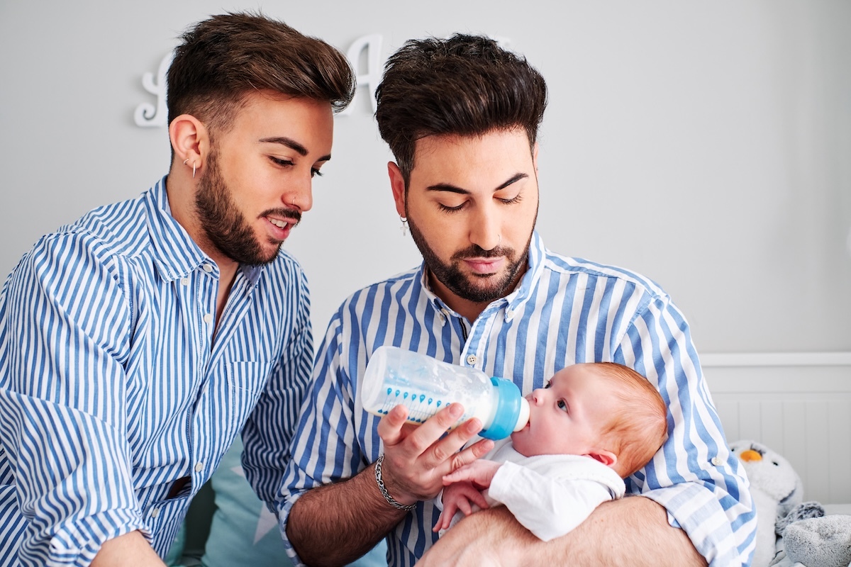 Two fathers bottle-feeding their newborn baby while sitting together in a nursery, both wearing matching blue striped shirts.