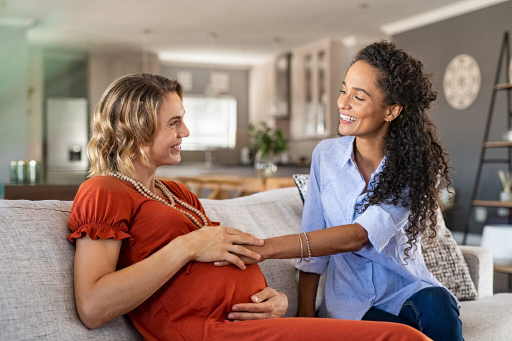 Two smiling women sitting on a couch in a cozy home setting. One woman is visibly pregnant, wearing an orange dress with beaded necklaces, and the other woman, wearing a light blue blouse and jeans, is gently touching her baby bump while they share a joyful moment together.