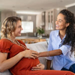Two smiling women sitting on a couch in a cozy home setting. One woman is visibly pregnant, wearing an orange dress with beaded necklaces, and the other woman, wearing a light blue blouse and jeans, is gently touching her baby bump while they share a joyful moment together.